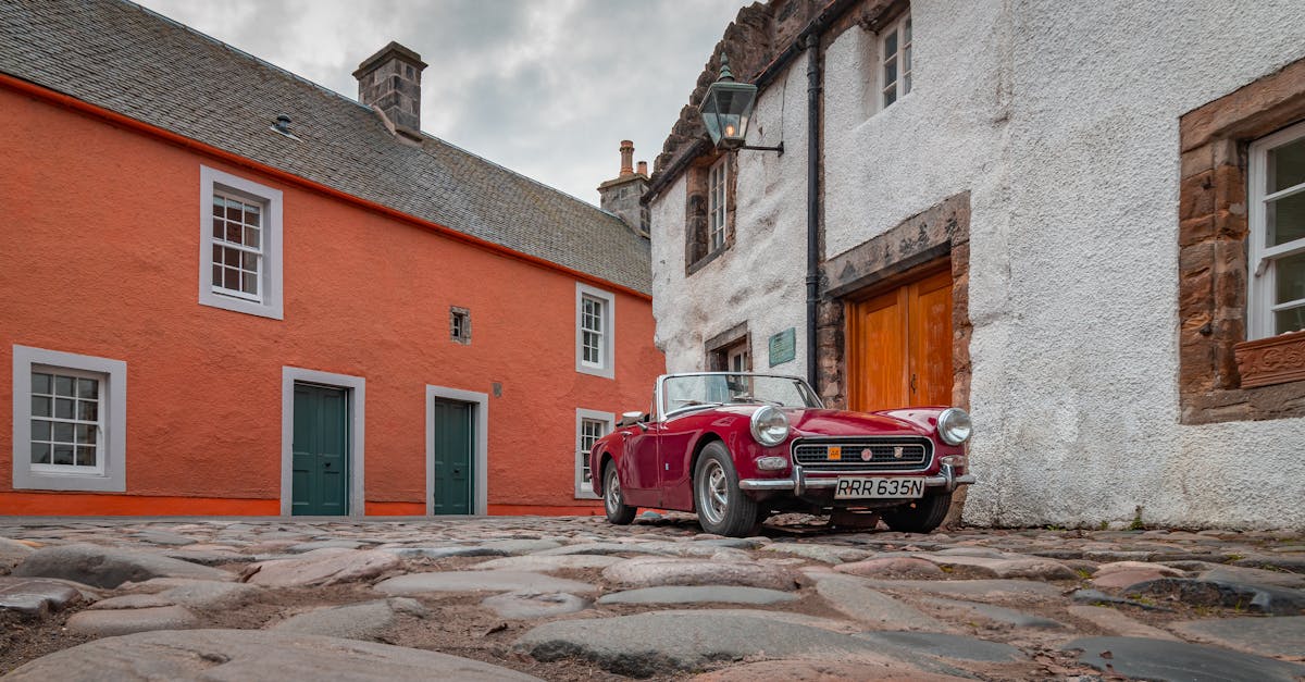 A classic vintage red convertible parked on cobblestone in Culross, Scotland, amidst historic buildings.