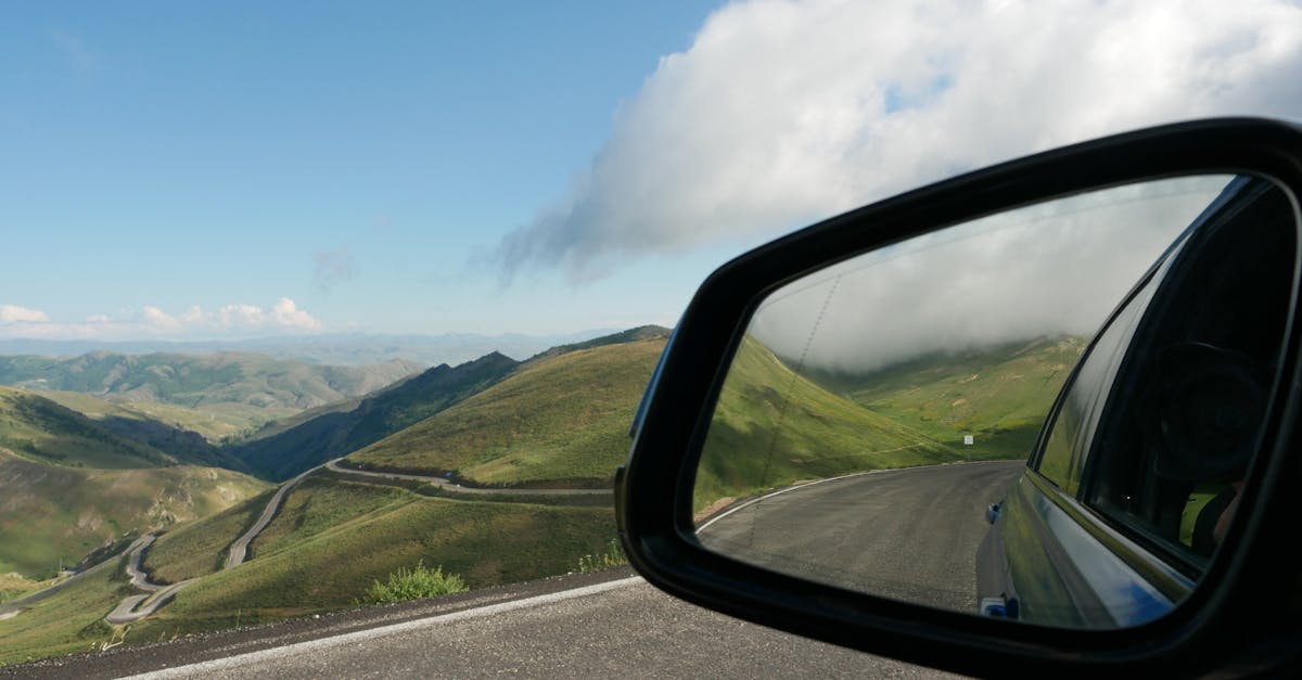 Rearview mirror reflecting winding road through lush hills under cloudy sky.