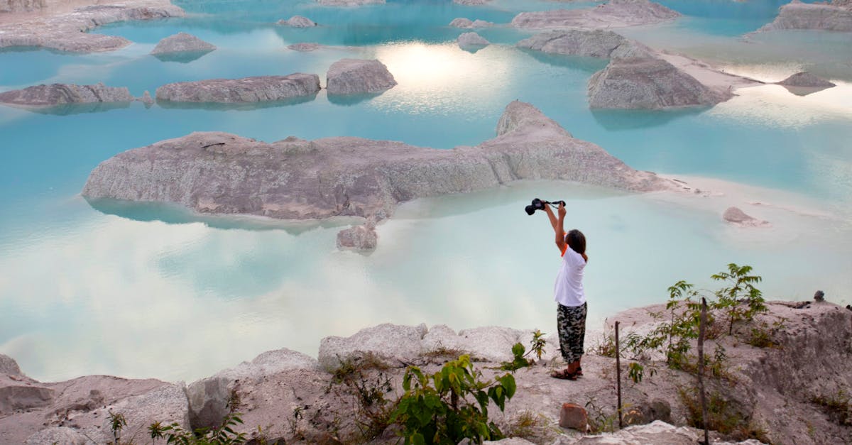 A woman takes a picture of a stunning blue lake amidst rocky terrain at sunrise.