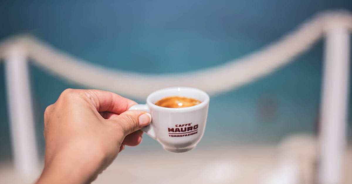 A hand holding an espresso cup over a blurred ocean background, capturing a serene coastal moment.