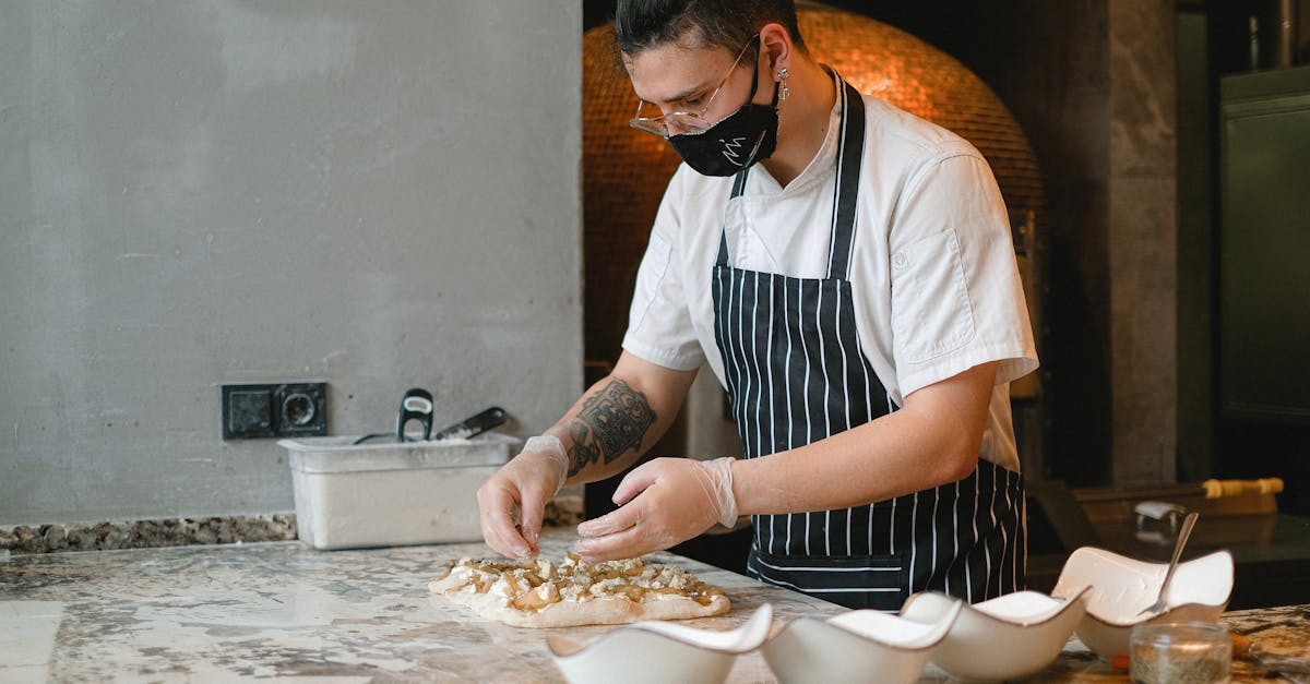 A chef wearing a face mask intricately prepares pizza dough in a professional kitchen.