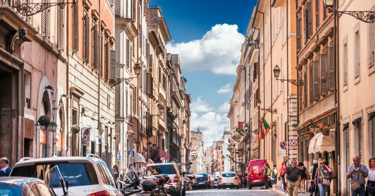 Narrow street with cars and motorcycles parked near historic buildings located in city in Europe and with people walking outside under blue sky in summer sunny day