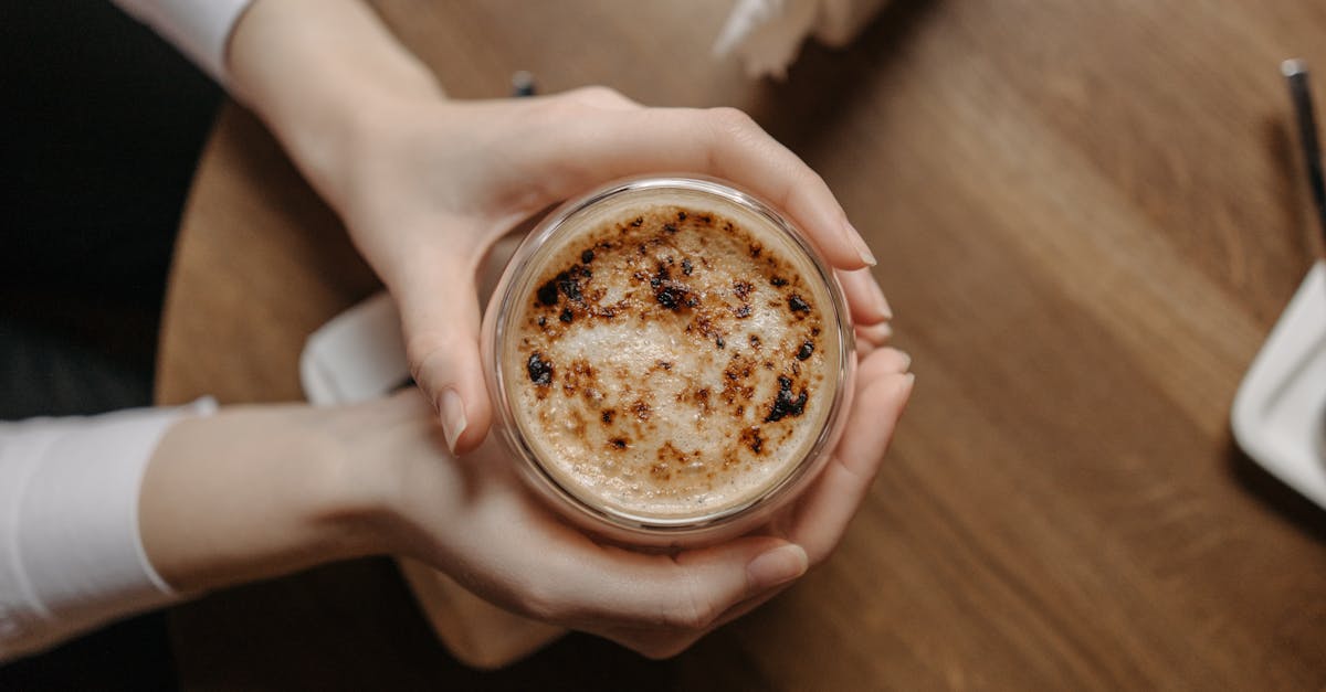 A top view of a cappuccino with foam in a glass held by hands over a wooden table.