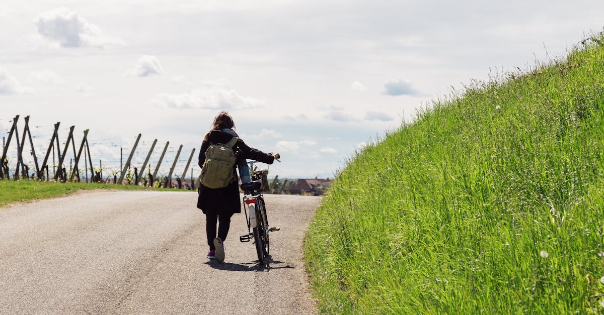 Woman walking with bicycle along a scenic country road with lush greenery and blue skies.