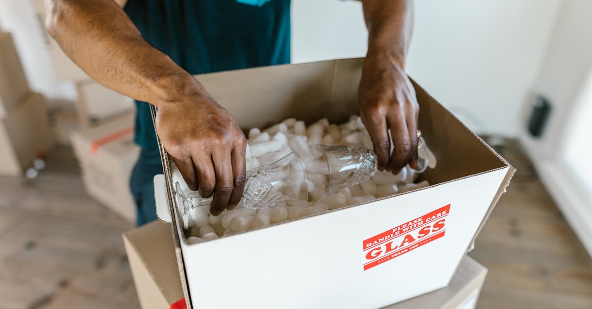 Hands carefully packing glassware into a cardboard box with padding for safe transport.