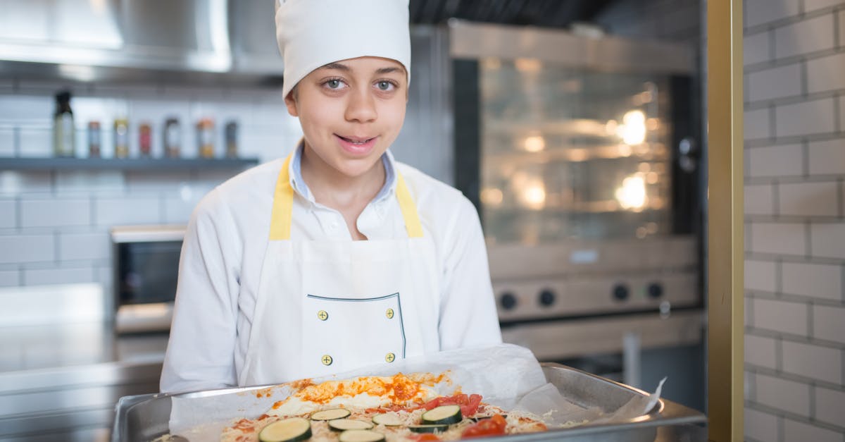 A young boy wearing a chef outfit prepares a pizza in a professional kitchen setting.