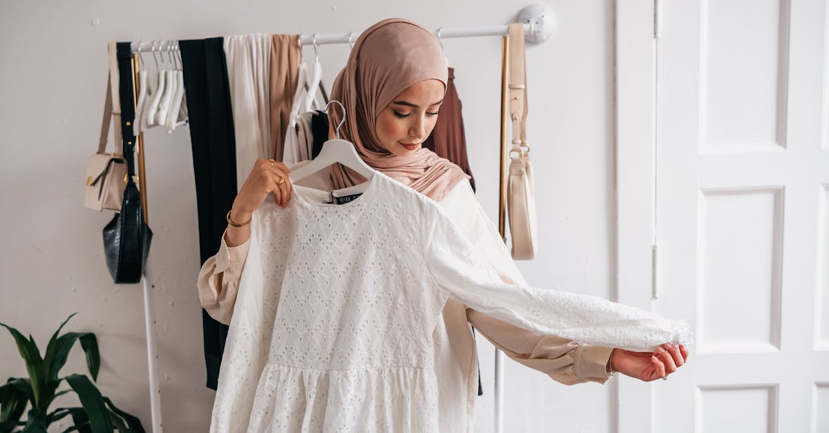 A Muslim woman in a hijab examines a white dress in a clothing boutique with racks of clothes.
