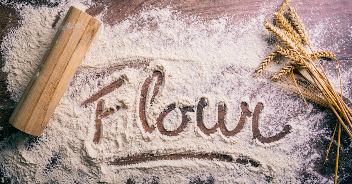 Flour scattered on a wooden table spelling 'flour,' with a rolling pin and wheat stalks nearby.